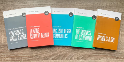 The Business of UX Writing, Design Is a Job, Inclusive Design Communities, Leading Content Design, and You Should Write a Book paperback books spread out on a dark wood table.