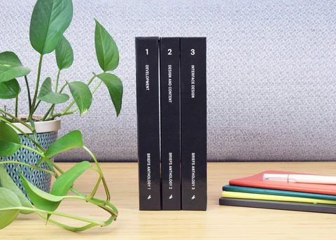 Three glossy hardcover black books standing on a light wood desk, between a green plant on the left and multicolored notebooks and a white pen on the right.