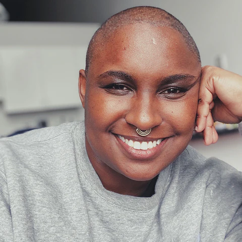 A Black nonbinary person smiles and leans against their hand. They are wearing a grey shirt and silver nose jewelry.