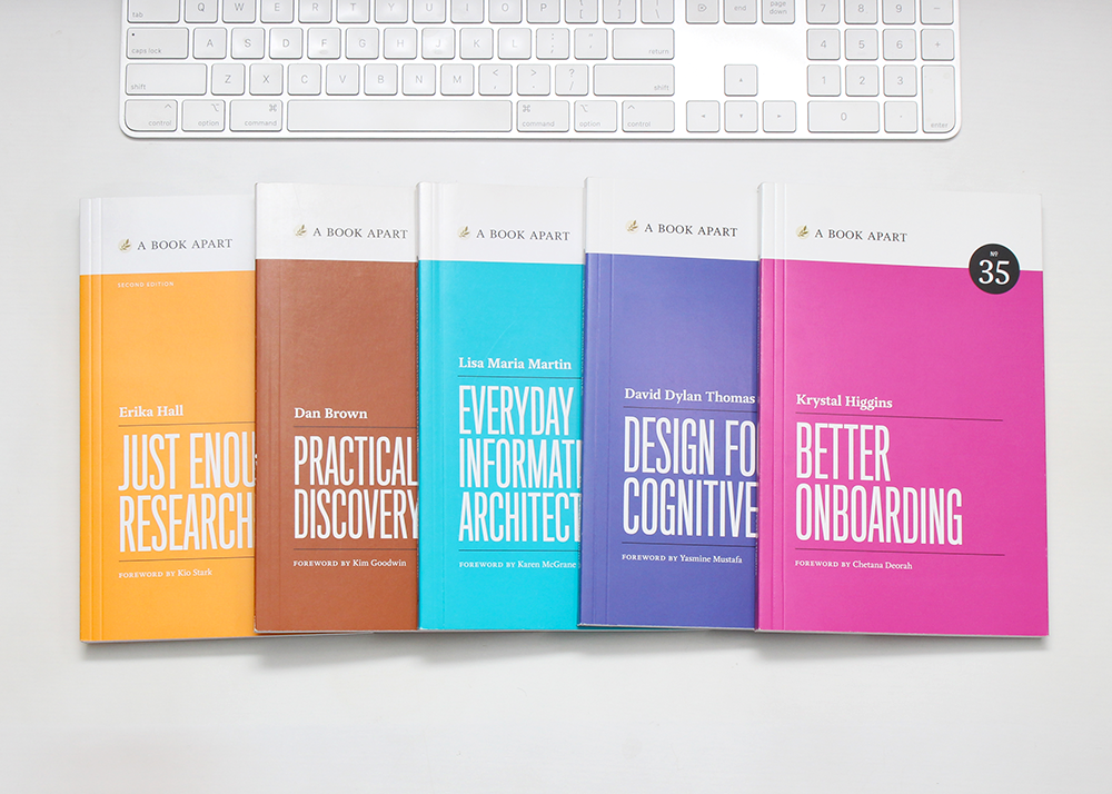 Just Enough Research, Practical Design Discovery, Everyday Information Architecture, Design for Cognitive Bias, and Better Onboarding paperbacks spread out on a white desk below a white keyboard.