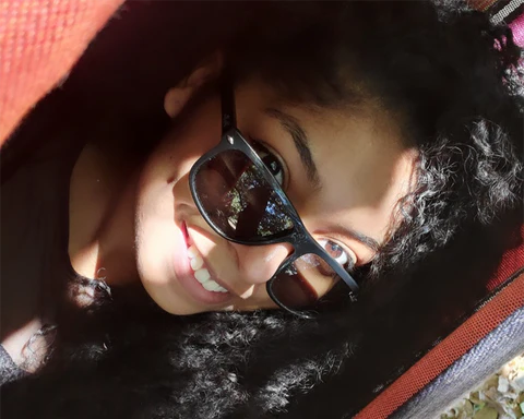 A woman with brown eyes and curly hair smiles and wears sunglasses.