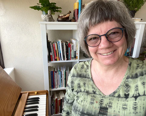 A white woman wearing glasses, sitting next to a piano, in front of a bookcase.